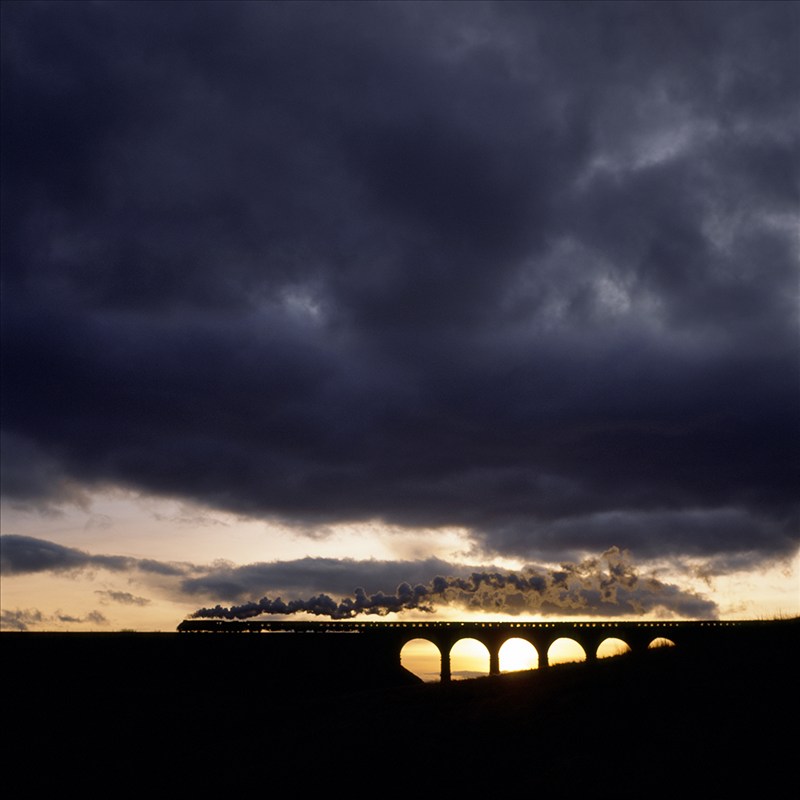 Classic train and classic architecture. The iconic Ribblehead Viaduct on the Settle and Carlisle railway line is crossed by an A4 steam locomotive on a charter. The entire scene is dwarfed by the massive sky at sunset.