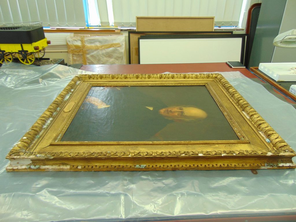 A portrait in a damaged gilt picture frame on the conservation workshop bench