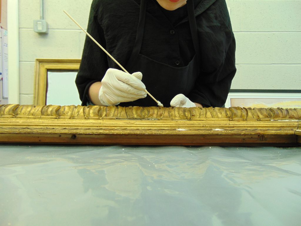 A conservator in black top and white latex gloves uses a cotton swab to clean a gilt picture frame