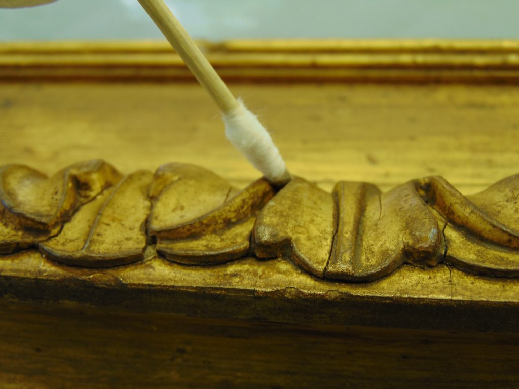 A close-up of a gilt picture frame, with a cotton swab being used to clean the carved design