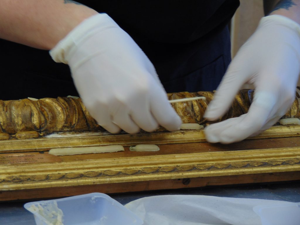 The white-gloved hands of a conservator applying replacement details to a gilt picture frame