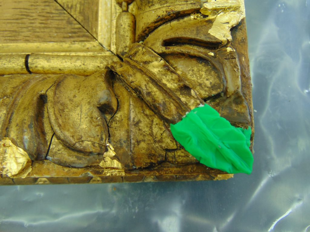 The corner of a gilt picture frame with a leaf detail added in green plasticine
