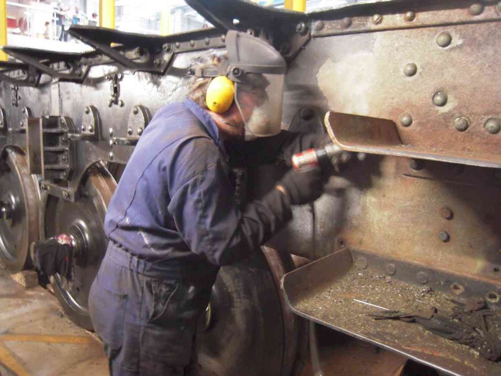 A man in coveralls and visor needle-gunning the locomotive's frames