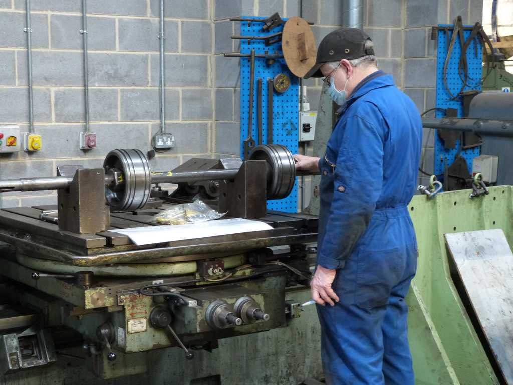 A white man wearing blue overalls, a face mask and black cap stands in the workshop