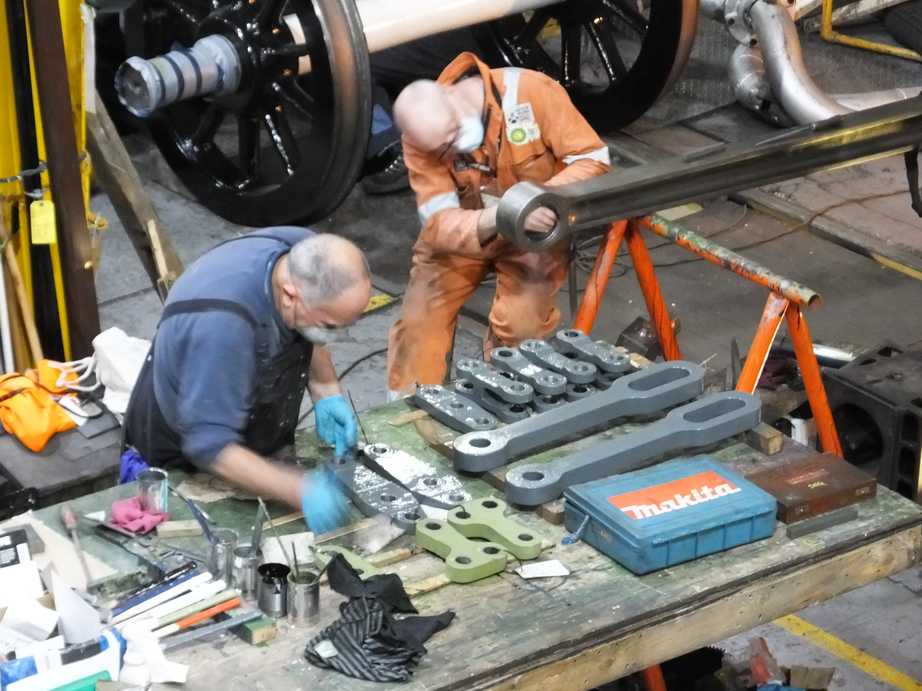 Two people in coveralls and face masks. One stands at a workbench while the other leans over a loco part.