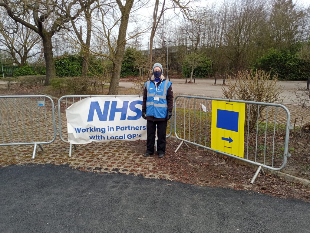 A woman in high-visibility vest and face mask stands by some temporary fencing. A banner on the fence reads 'NHS, working in partnership with local GPs'
