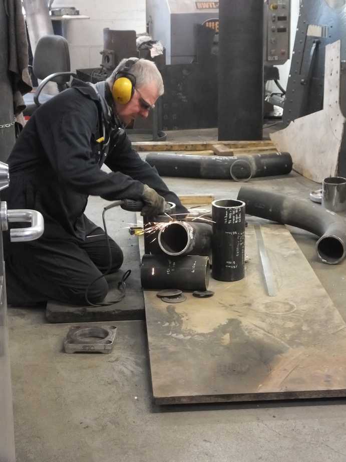 A man wearing yellow ear defenders kneels on the wrokshop floor, working on some pieces of pipin
