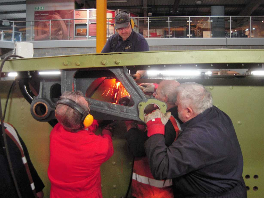 It gets busy around a hot rivet when there's an expansion link bracket in the way. Billy in the red overalls puts the rivet in the hole with tongs, Pete knocks it in with a hammer, Trevor moves the jamback and then Mick can close the rivet.