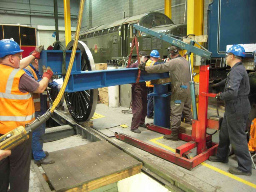 Lifting beam being inserted between the tank and frames by a team of people in overalls and hard hats.