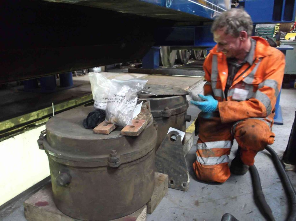 Man in orange high-visibility overalls kneeling by locomotive brake cylinders.