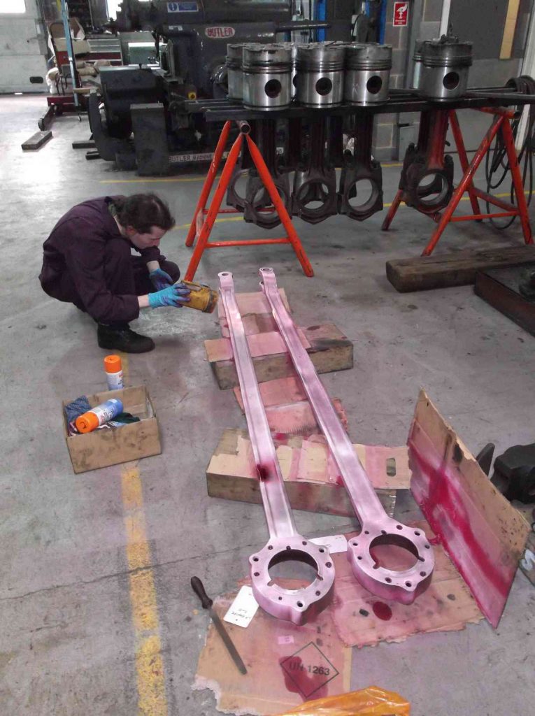 Two return crank rods laid out on the workshop floor as a team member inspects for defects
