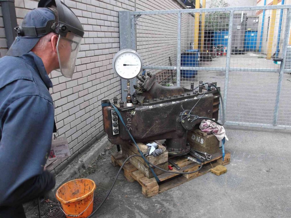 Man in a plastic face visor stands by the superheater header, outside the workshop