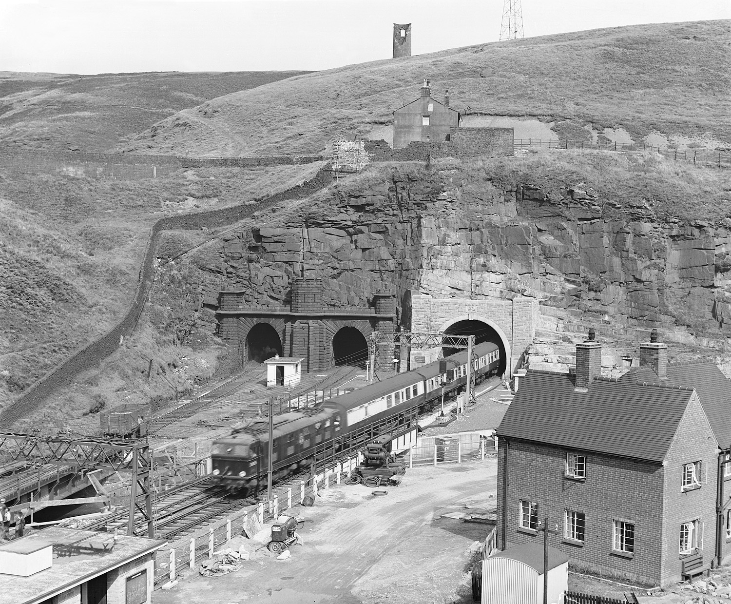 A passenger train hauled by an unidentified EM-type BO-BO electric locomotive emerges from Woodhead Tunnel in 1954. The electrification scheme was delayed by the Second World War and included the construction of a new tunnel at Woodhead. The old tunnels are to the left, which were taken out of use.